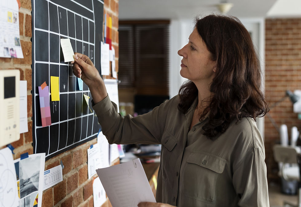 Woman places sticky note on a black grid board in office, surrounded by papers. She's focused, wearing a gray shirt, brick wall background.