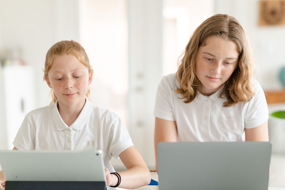 Two young students in white uniforms work on tablets and laptops at a desk in a bright room, focused and engaged.